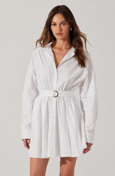 A woman is wearing a white, long-sleeved shirt dress with a collar and button-down front. The dress features a gathered waist with a matching fabric belt that has a silver buckle. The skirt portion of the dress is pleated, creating a slightly flared silhouette. The woman's hair is long and wavy, and she has a neutral expression on her face.